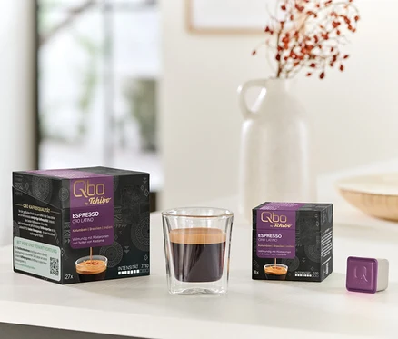 Auf einem Tisch stehen Qbo Espresso ORO TOLIMA Kapselpackungen (27 und 8 Kapseln) neben einem Glas Espresso und einer einzelnen Kapsel.