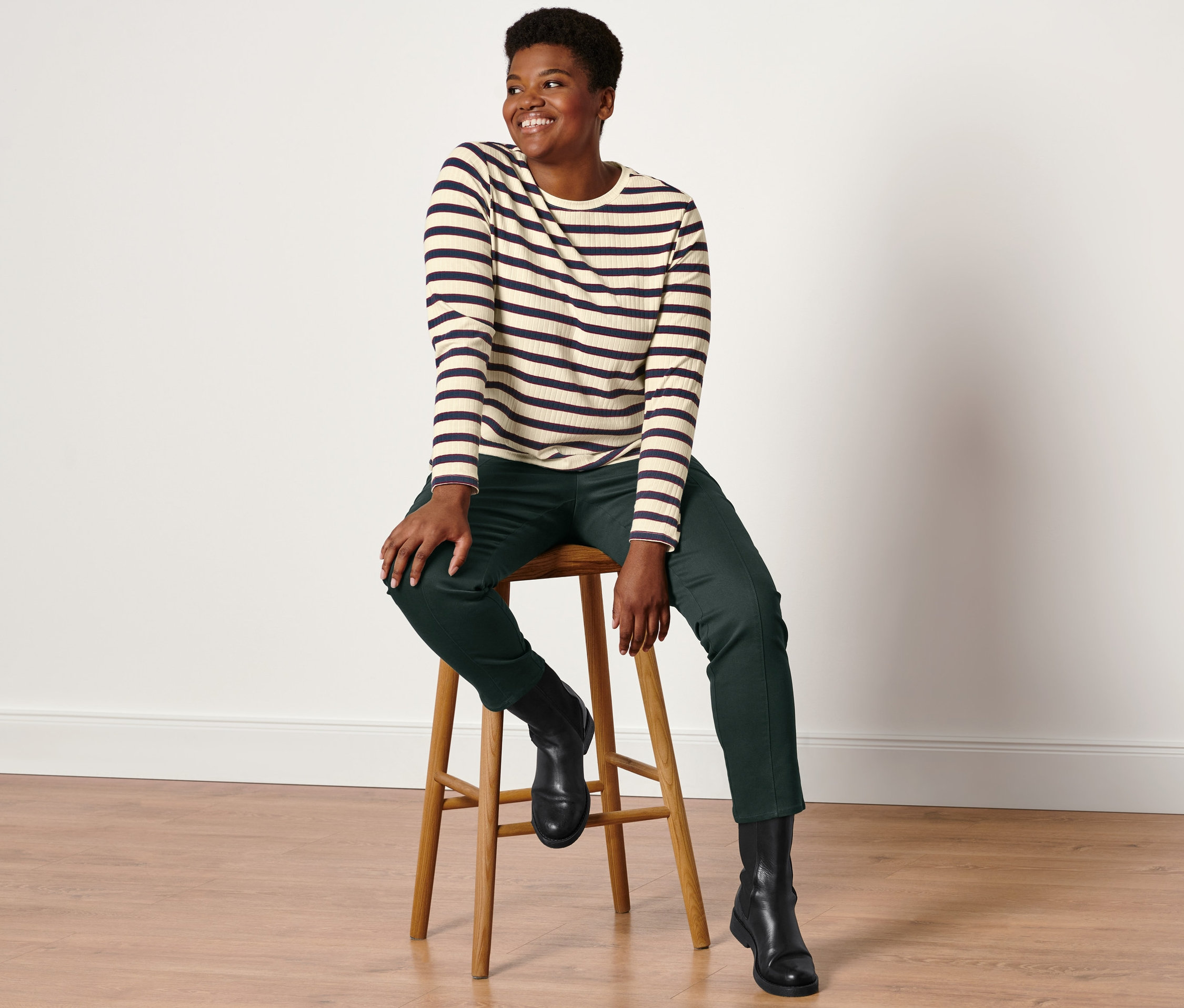 Frau sitzt auf einem Hocker und trägt ein geripptes Langarmshirt, eine Twillhose mit Komfortbund in Dunkelgrün und Lederboots.