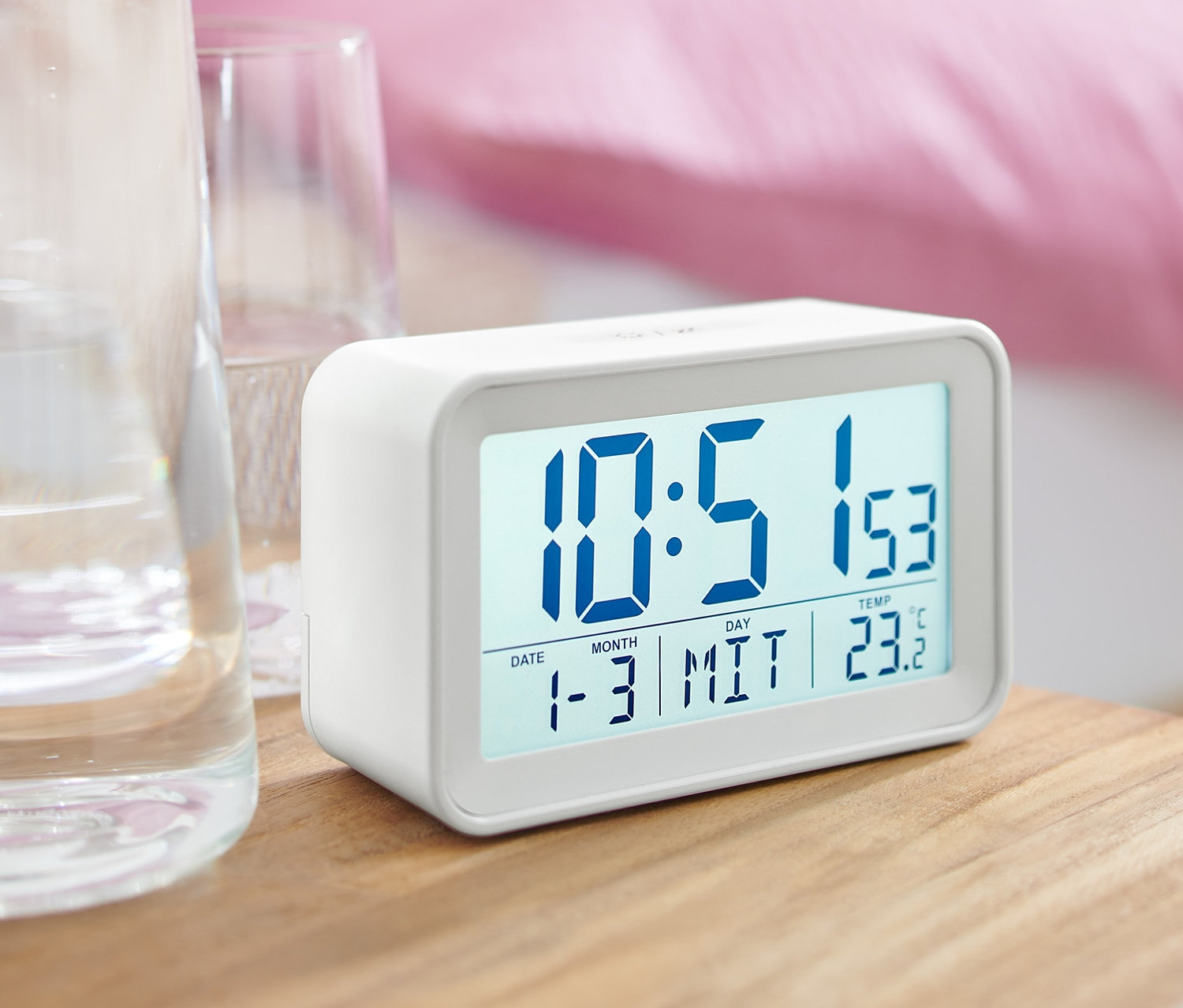 Ein weißer digitaler LCD-Wecker steht auf einem Nachttisch neben einem Glas Wasser.