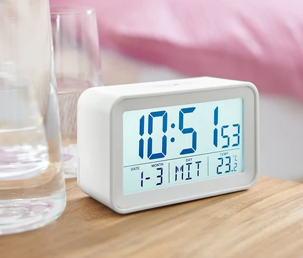 Ein weißer digitaler LCD-Wecker steht auf einem Nachttisch neben einem Glas Wasser.