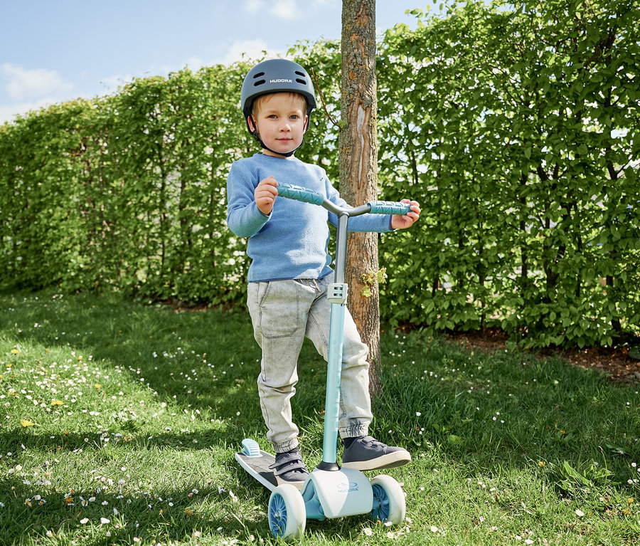Junge mit Helm steht auf blauem HUDORA Tri-Scooter auf einer Wiese.