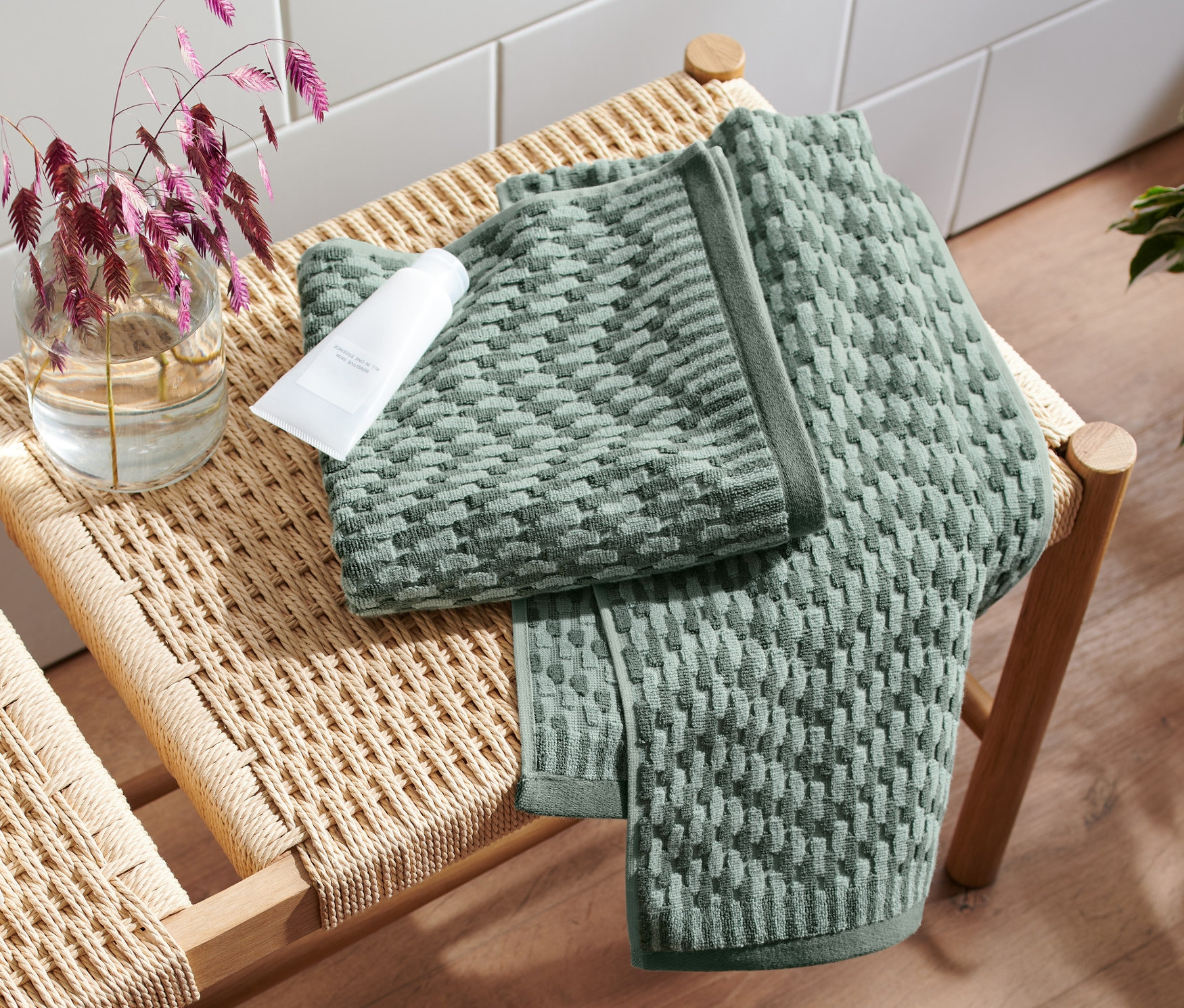 Auf einer Weidenbank liegen zwei grün-salbeifarbene Jacquard-Handtücher und eine weiße Cremetube neben einer Vase mit lila Blumen.