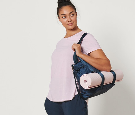 Frau trägt Yogamatte in einer Tasche über der Schulter, dazu eine Yogahose und ein hellrosa Longshirt.
