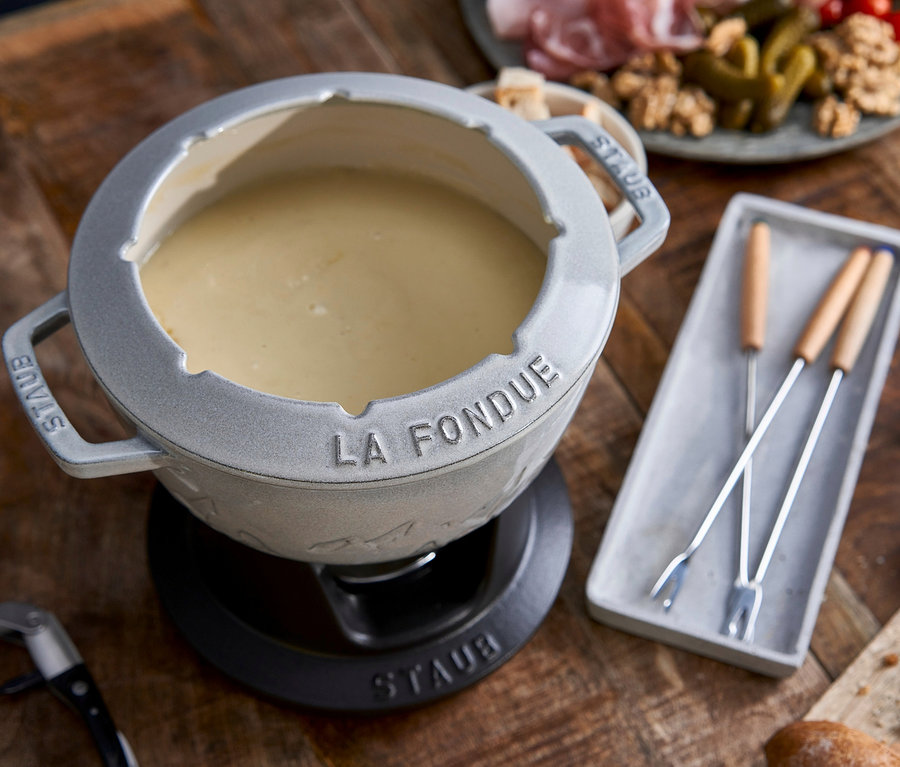 Auf einem Holztisch steht ein weißes STAUB Fondue-Set mit Käsesauce, daneben liegen Fonduegabeln, Brot, Wein und ein Teller mit Wurstwaren.