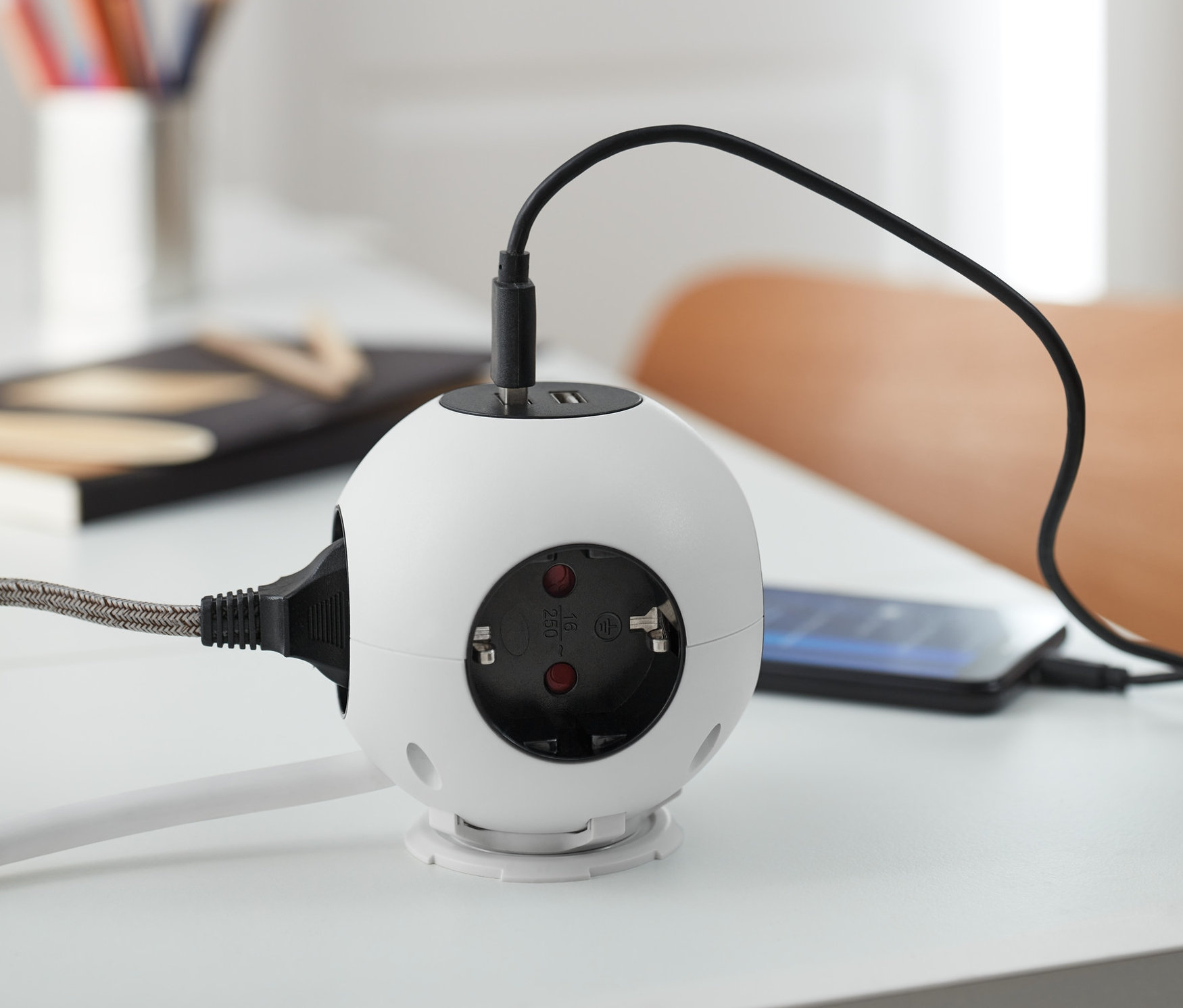 Ein weißer Steckdosenball mit USB-Ladefunktion steht auf einem weißen Tisch und lädt ein Mobiltelefon.