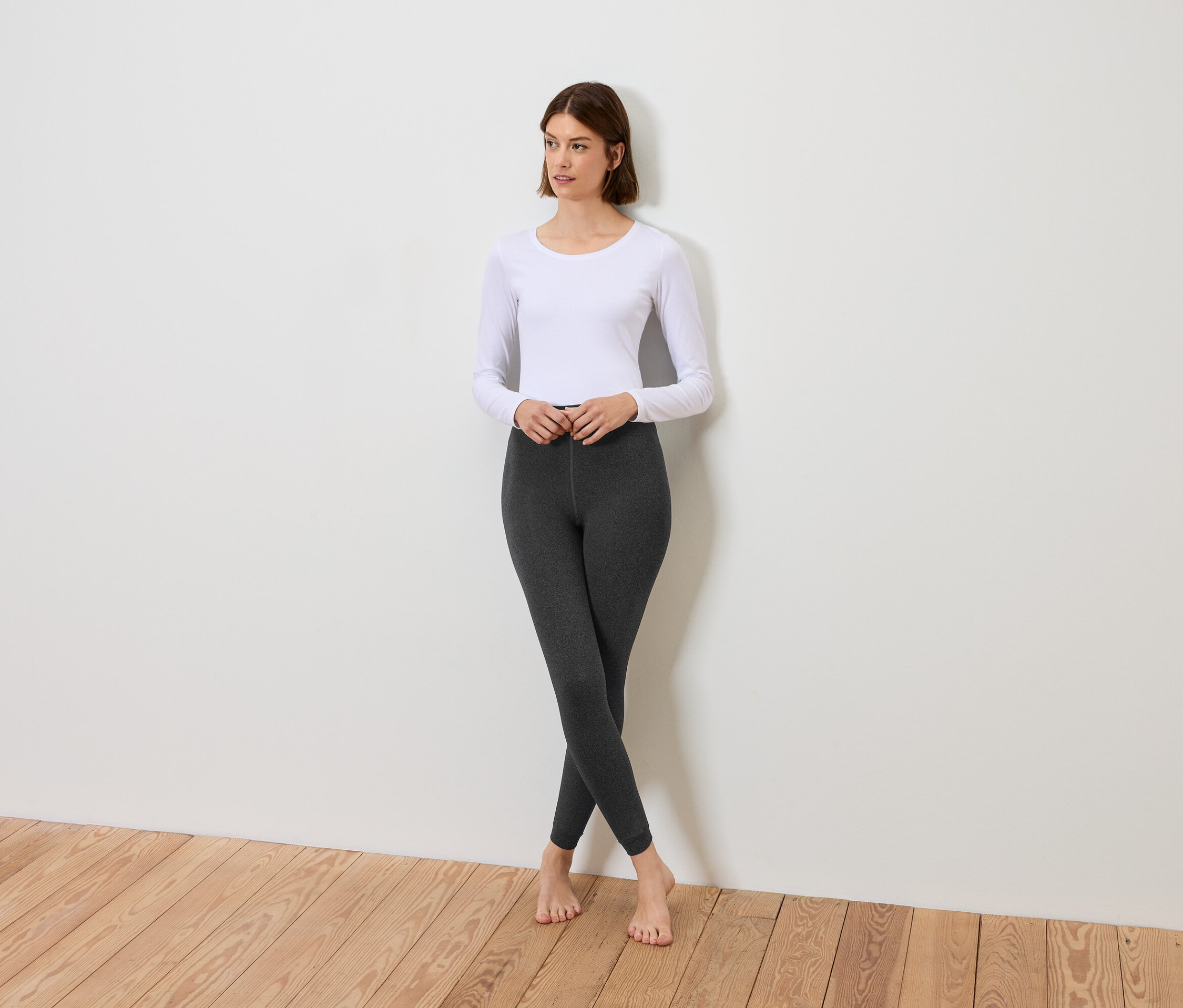 Eine Frau steht mit gekreuzten Beinen, bekleidet mit einem weißen Langarmshirt und grauen Leggings.