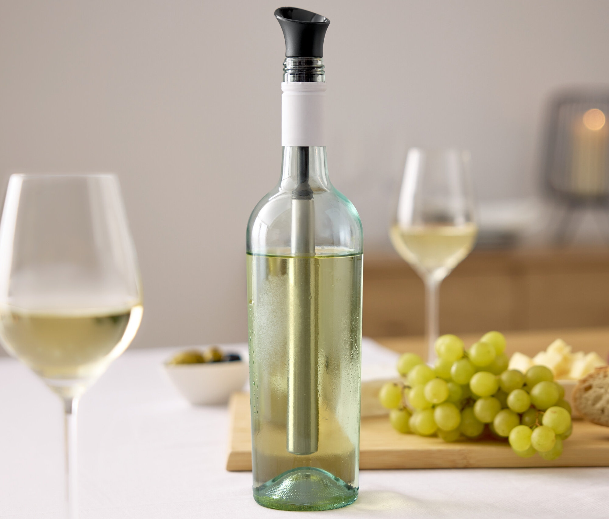 Weinflasche mit Wein-Kühlstab, zwei Gläser Wein, Trauben, Käse und Brot auf einem Tisch.