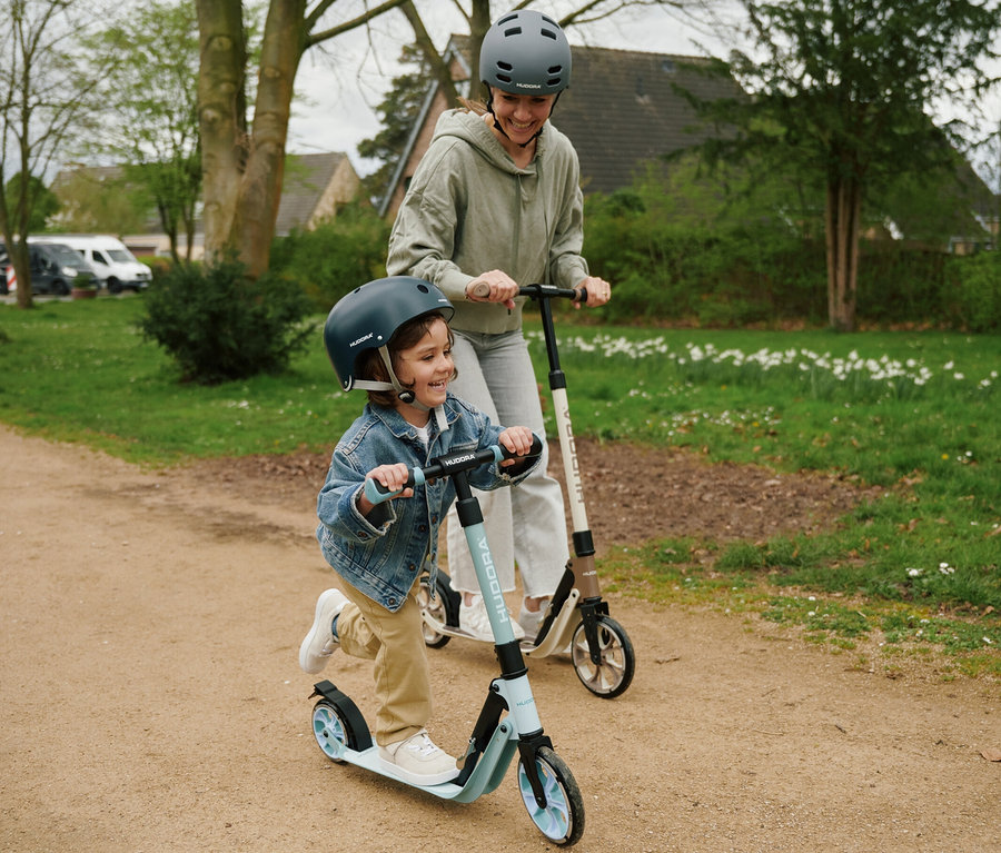 Ein Kind und ein Erwachsener lachen und fahren mit HUDORA BigWheel 180 Advanced Junior Rollern auf einem Feldweg.