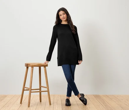 Frau mit schwarzem Pullover und Jeans steht neben einem Holzhocker.