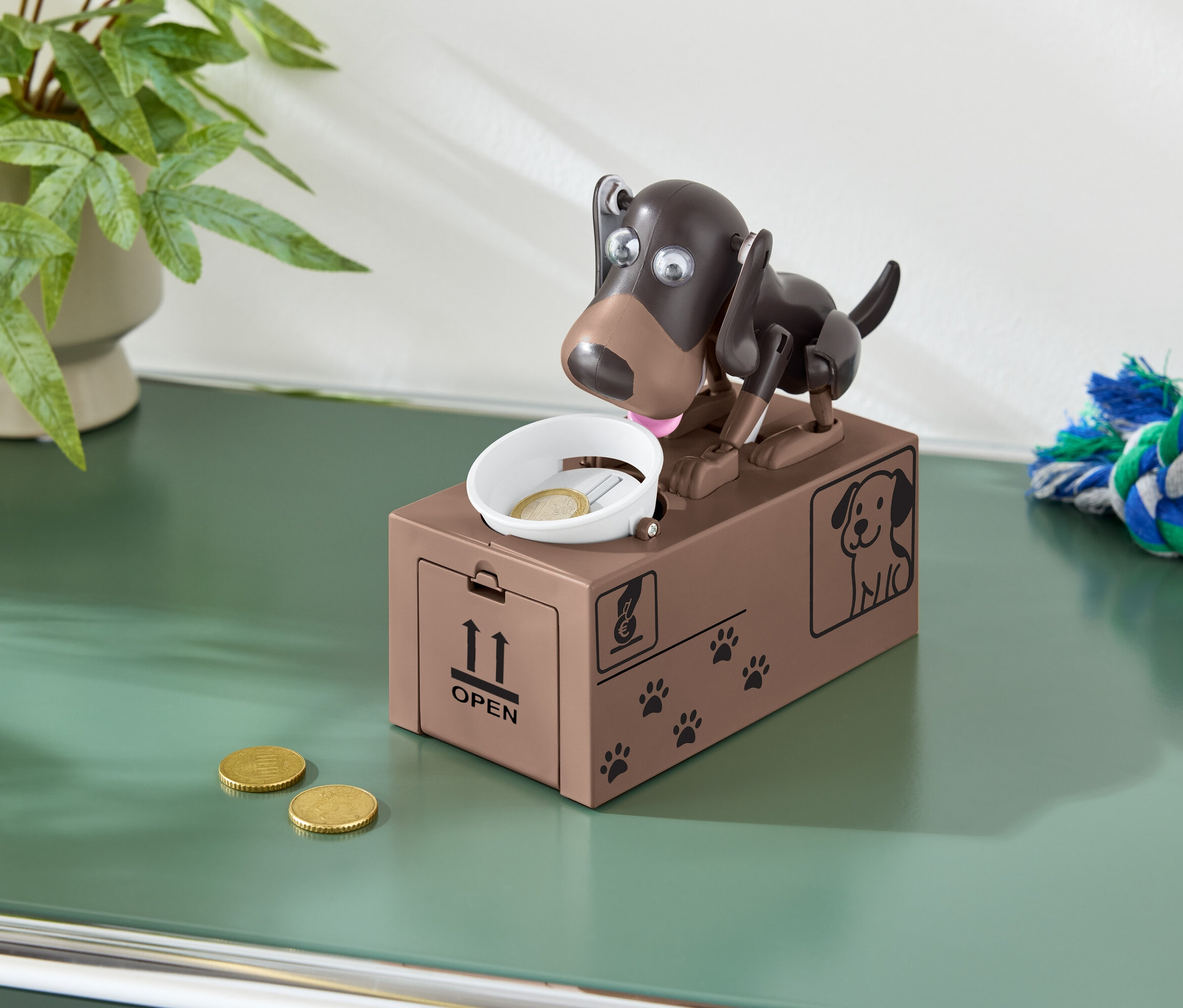 Braune Spardose in Hundeform mit einer Münze in einer Schale und zwei Münzen auf einem grünen Tisch.