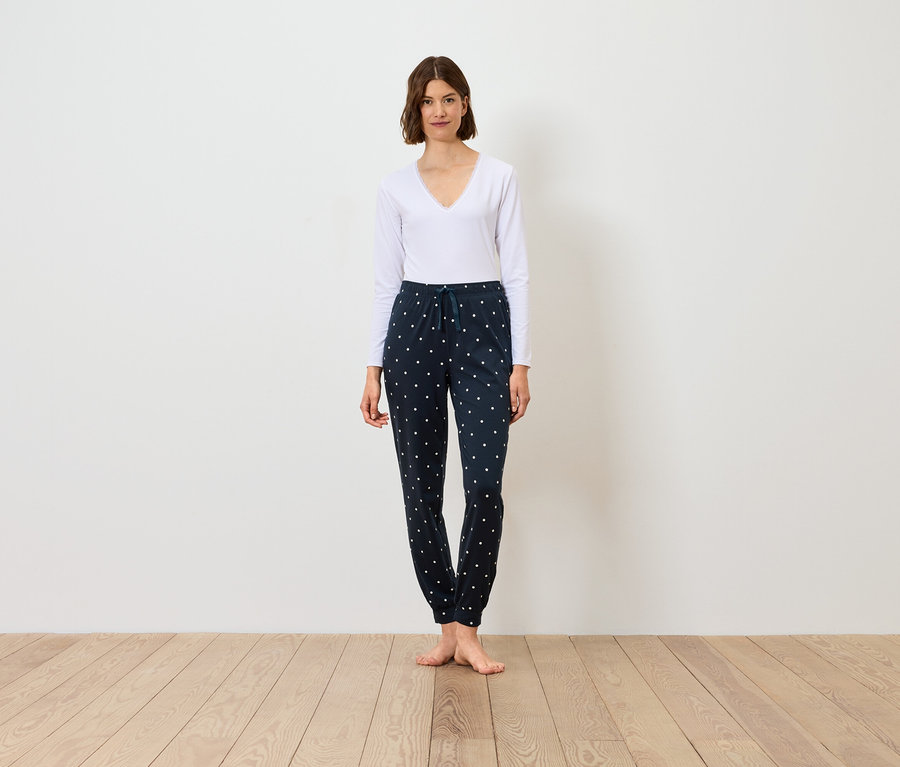 Frau steht und trägt ein weißes Langarmshirt und eine dunkelblaue Hose mit Muster.