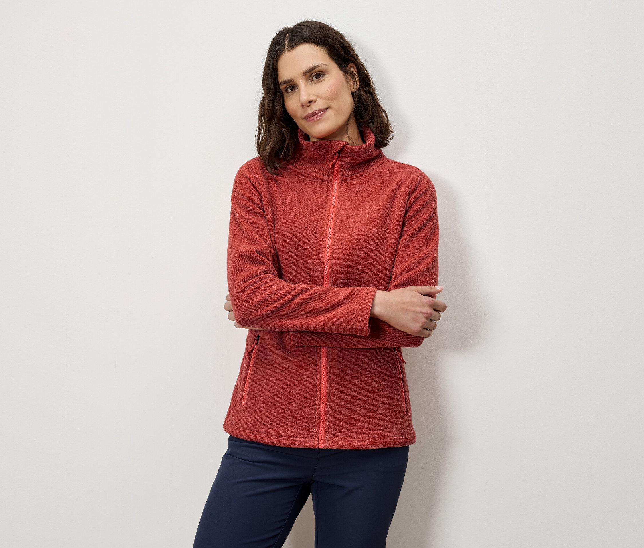 Frau posiert mit verschränkten Armen in orangefarbener Fleecejacke und marineblauer Hose.