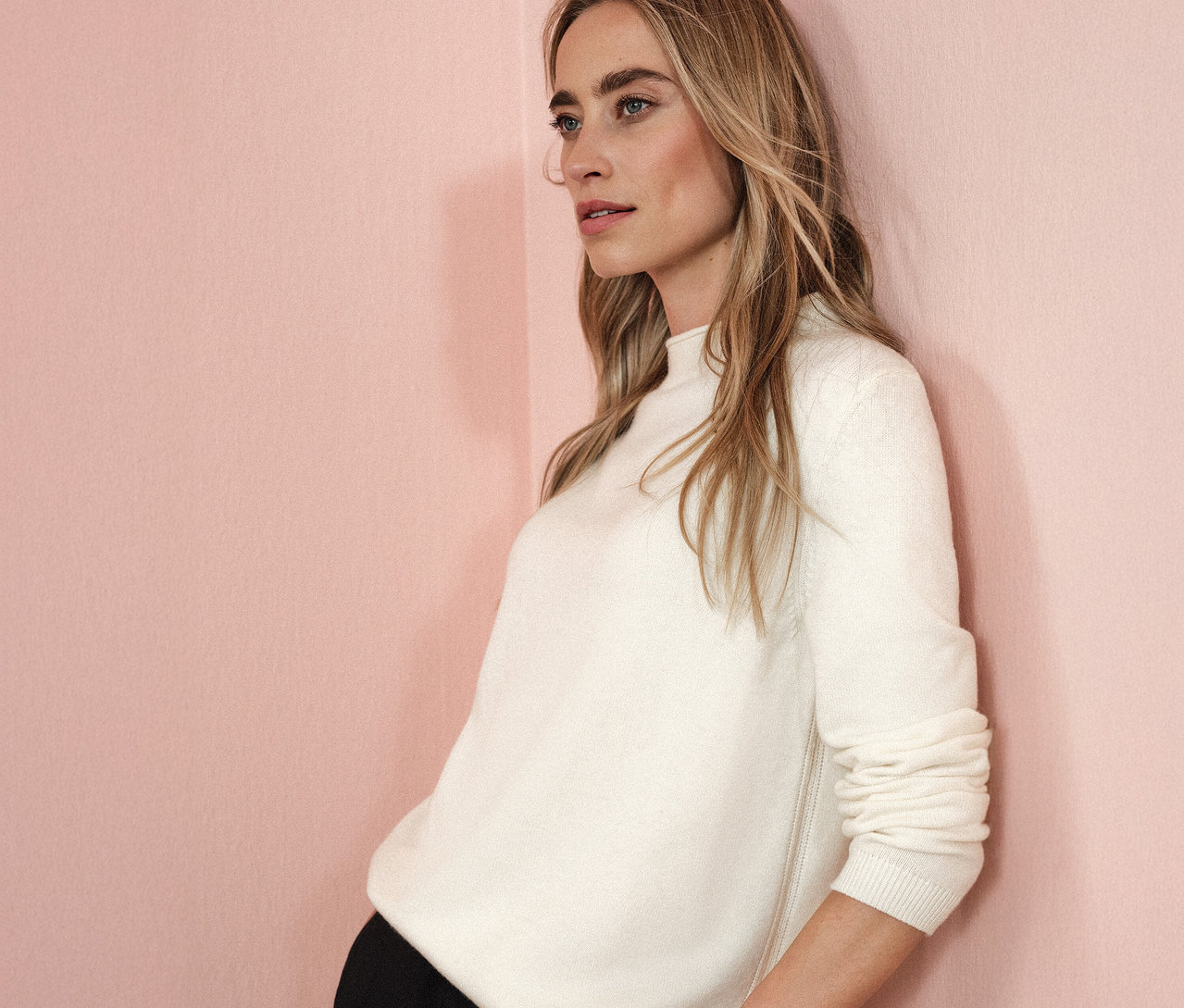 Frau lehnt an einer rosa Wand und trägt einen beigen Street One Strickpullover mit Rollkante.