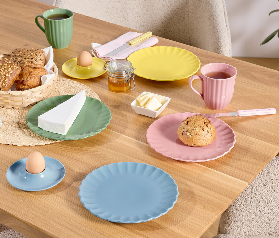 Ein Holztisch ist mit bunten Tellern, Tassen und Schüsseln mit Speisen gedeckt, darunter Brot in einem Korb, ein Ei in einem Eierbecher und ein Stück Käse.