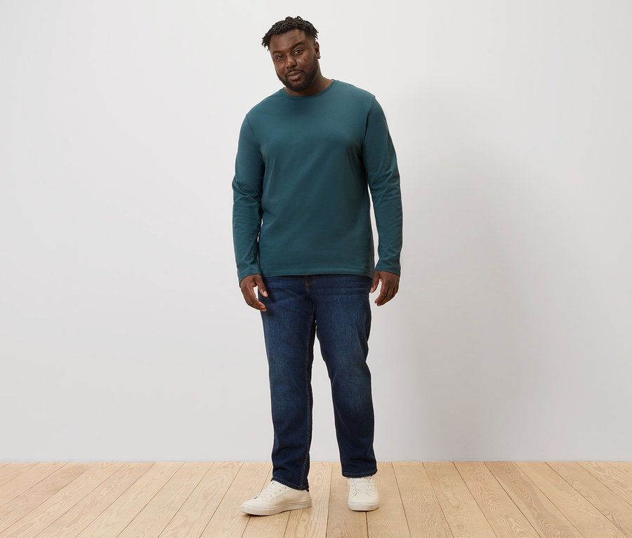 Mann steht und trägt Jeans und ein grünes 2 Langarmshirts.