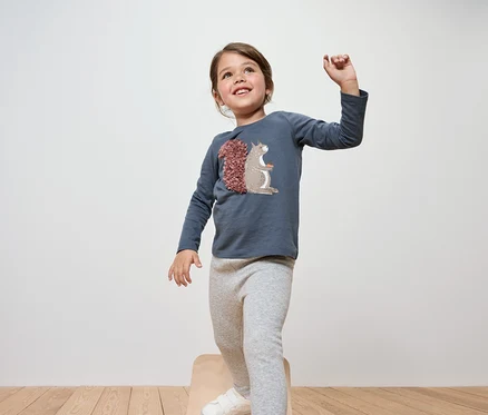 Ein lächelndes Mädchen steht auf einem Holzhocker und trägt ein blaues Langarmshirt und graue Leggings.
