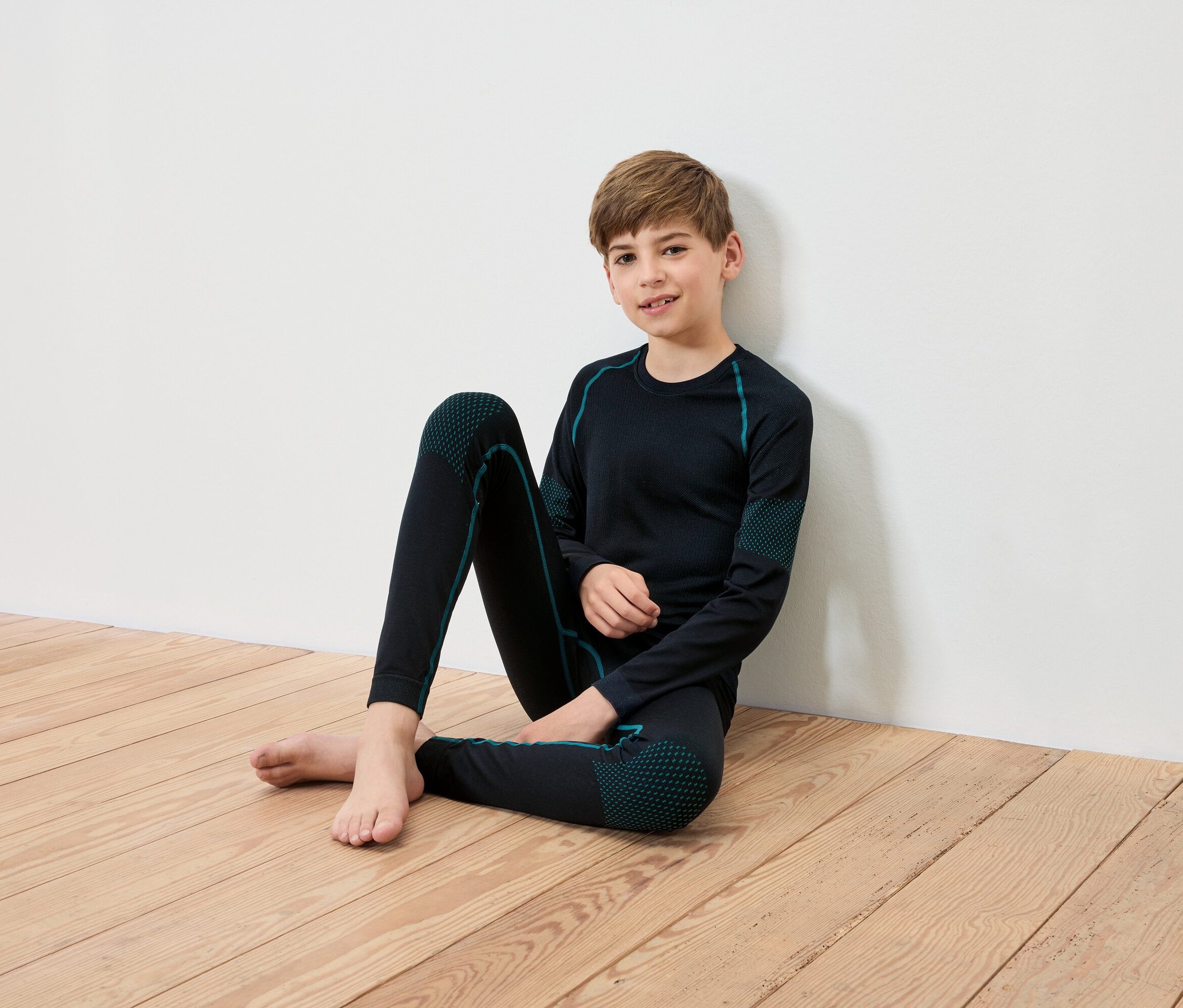 Ein Junge sitzt auf einem Holzboden und lehnt an einer weißen Wand. Er trägt schwarze Thermounterwäsche mit türkisen Details.