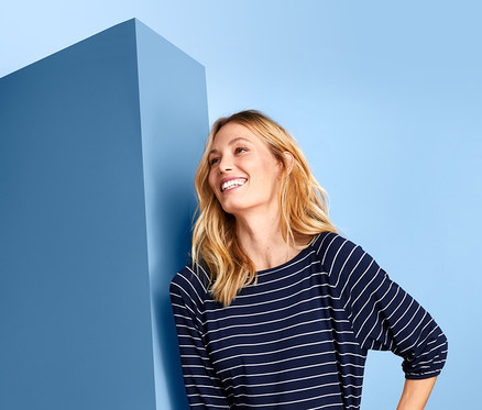 Eine Frau lächelt und lehnt an einer blauen Wand, sie trägt ein Shirt mit Fledermausärmeln und eine weiße Hose.