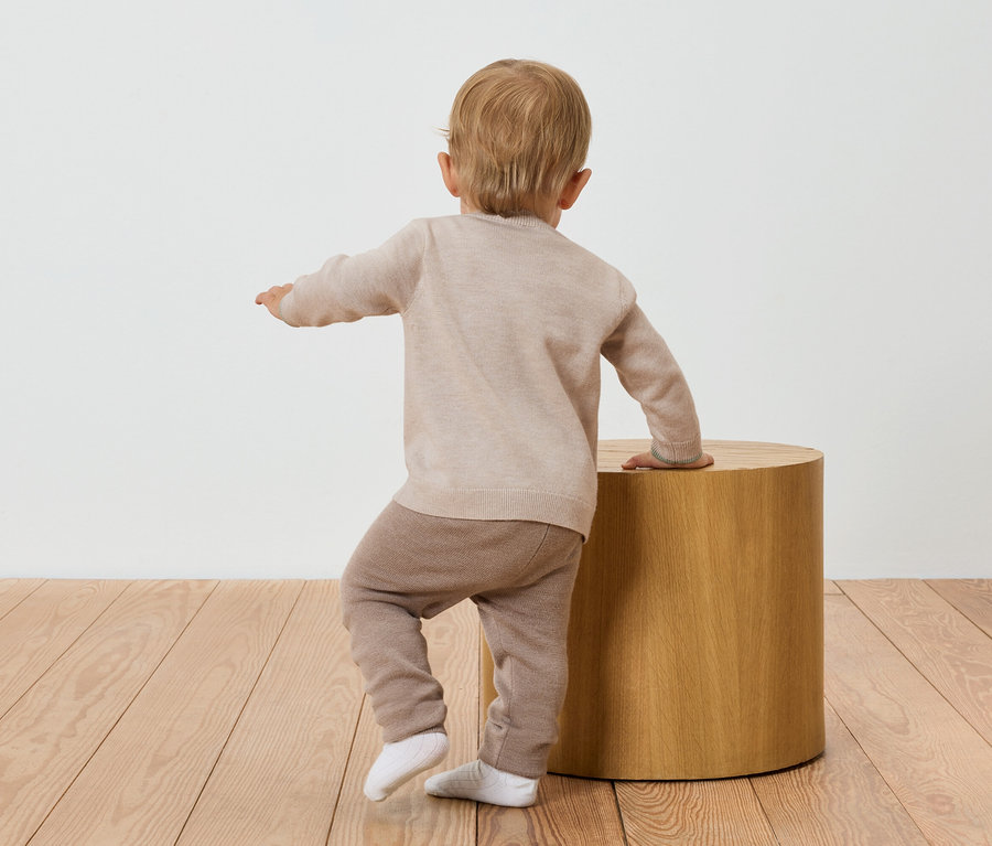 Kleinkind in beige Pullover und brauner Hose steht mit dem Rücken zur Kamera an einem Holztisch.