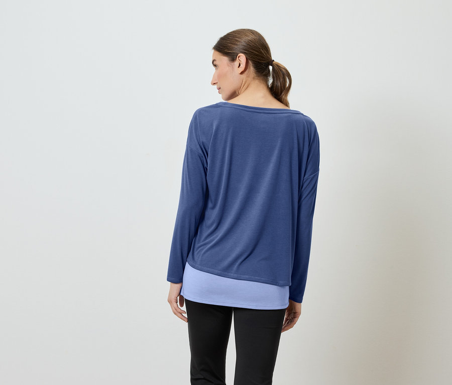 Frau mit dem Rücken zur Kamera trägt ein blaues Langarmshirt und eine schwarze Hose.