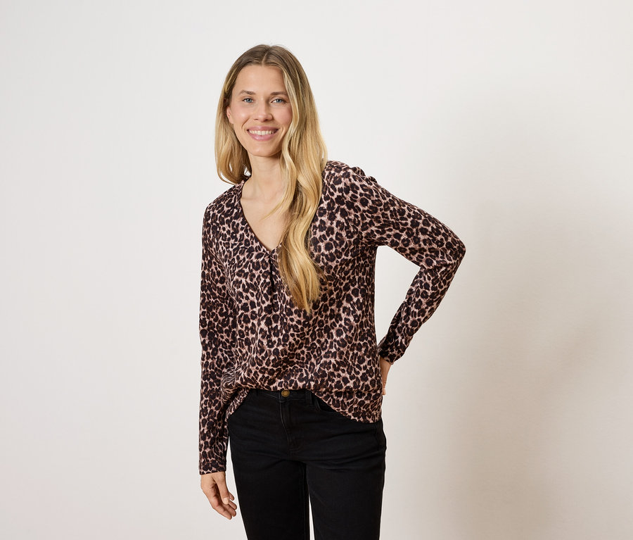 Frau mit langen blonden Haaren posiert in einer Bluse mit Tiermuster und schwarzen Hosen.