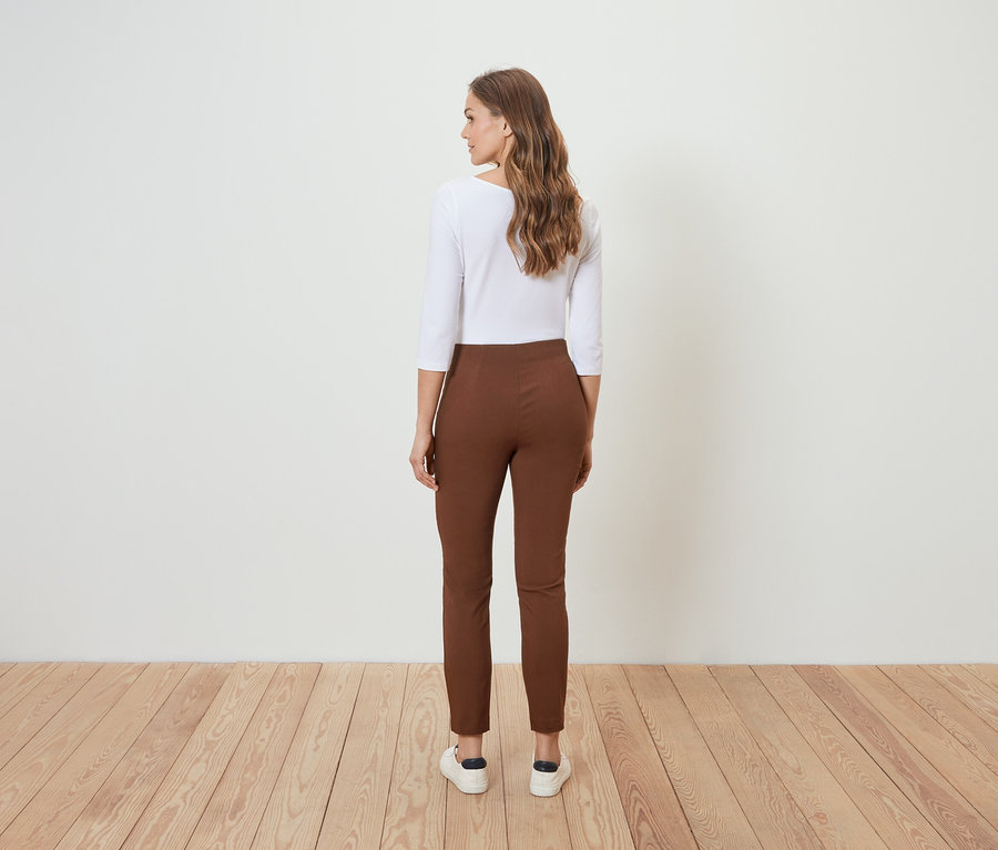 Eine Frau steht mit dem Rücken zur Kamera und trägt eine braune Hose und eine weiße Bluse.