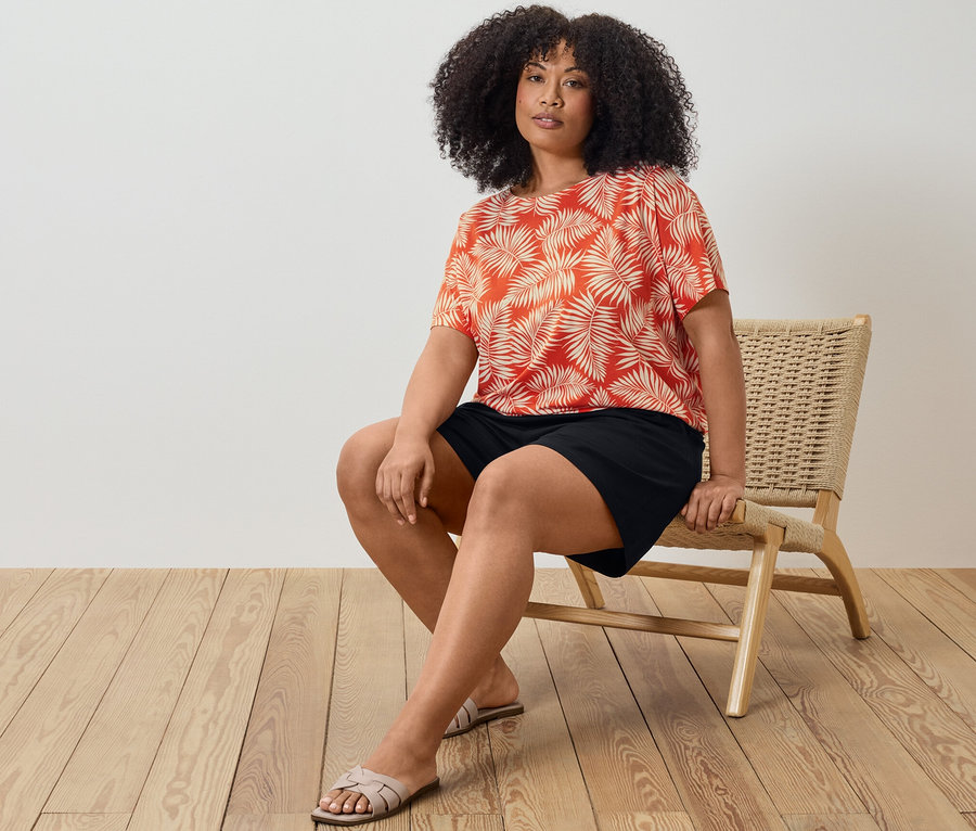 Eine Frau sitzt auf einem Stuhl und trägt ein bedrucktes Blusenshirt und schwarze Jerseyshorts.