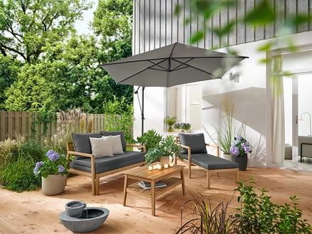 Auf einer Holzterrasse stehen ein ausziehbarer Gartensessel, ein ausziehbares Gartensofa, ein Garten-Couchtisch, ca. 80 x 50 cm, und ein Solar-Gartenbrunnen. Ein anthrazitfarbener Leco Ampelschirm spendet Schatten. 2 Outdoor-Kissen mit Leinen-Haptik und 6 LED-Outdoor-Akku-Teelichter dekorieren den Bereich.