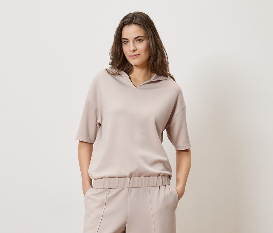 Frau mit braunen Haaren trägt ein beigefarbenes Kapuzenoberteil und eine passende Hose vor einem schlichten Hintergrund.