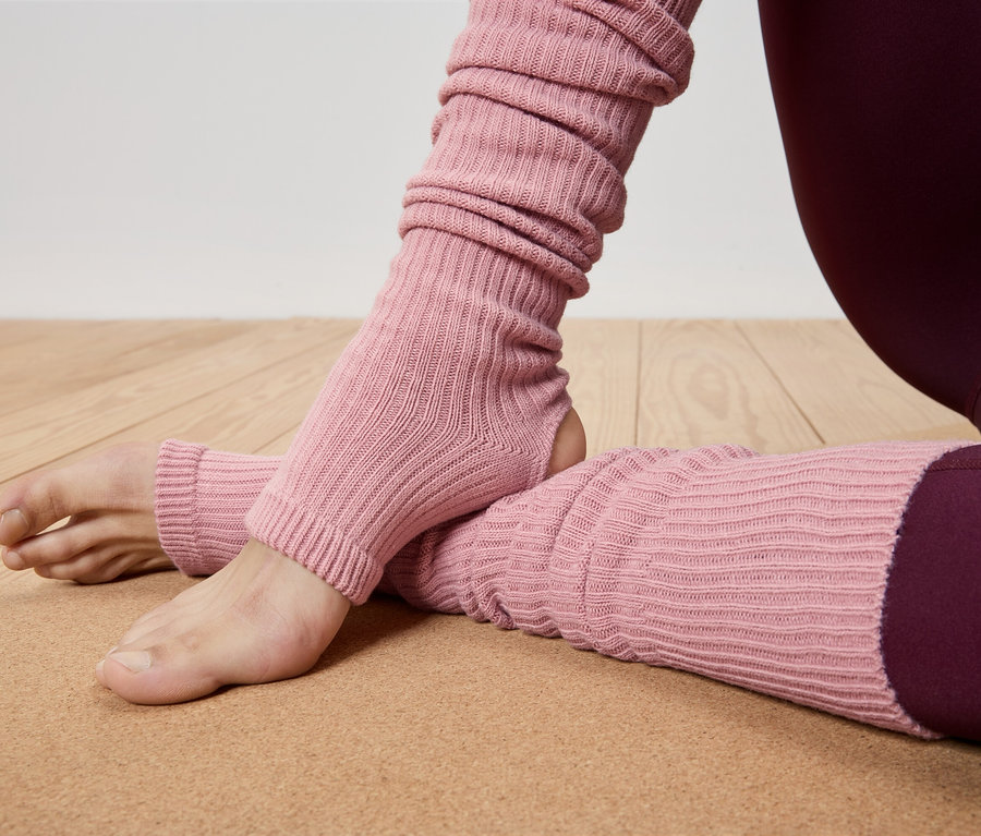 Nahaufnahme einer Person, die rosafarbene Beinstulpen und bordeauxfarbene Leggings auf einer braunen Yogamatte trägt.