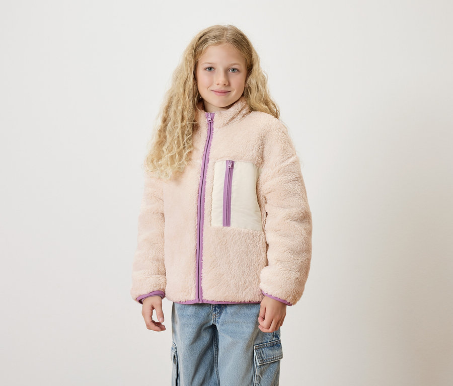Mädchen mit blonden, gelockten Haaren trägt eine cremefarbene Teddyjacke mit lila Akzenten und Jeans.