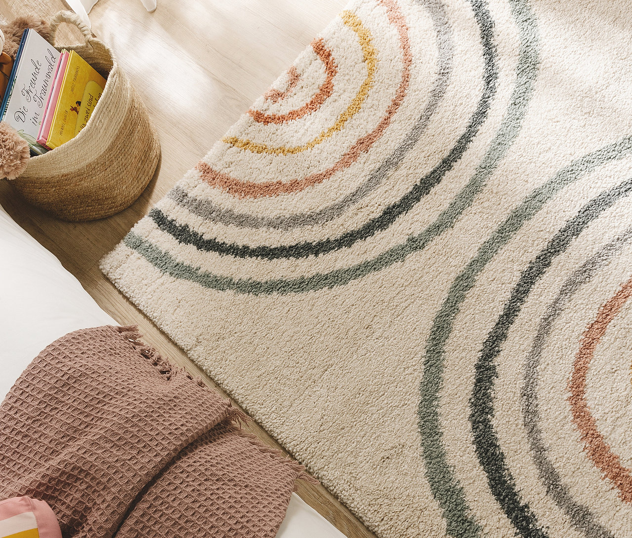 Beiger Teppich mit bunten, konzentrischen Halbkreisen auf Holzboden, daneben ein Korb mit Büchern und eine gefaltete, rosa Decke.