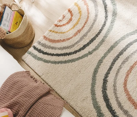 Beiger Teppich mit bunten, konzentrischen Halbkreisen auf Holzboden, daneben ein Korb mit Büchern und eine gefaltete, rosa Decke.