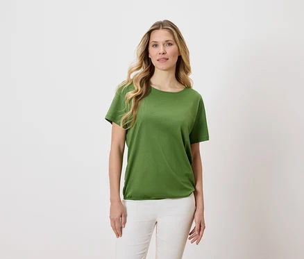 Frau mit blonden, gewellten Haaren trägt ein grünes T-Shirt und weiße Hosen vor einem hellen Hintergrund.
