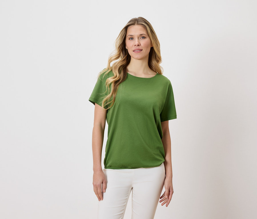 Frau mit blonden, gewellten Haaren trägt ein grünes T-Shirt und weiße Hosen vor einem hellen Hintergrund.