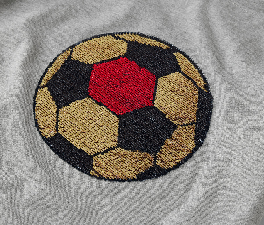 Auf einem grauen Stoff ist ein Fußball aufgenäht. Der Ball ist aus goldenen, schwarzen und roten Pailletten.
