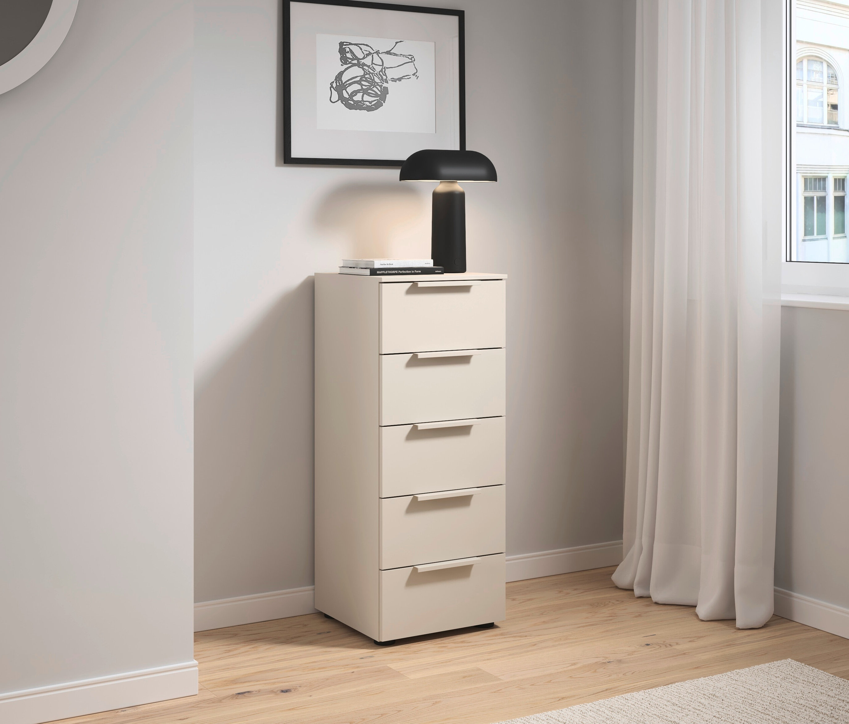Ein beiger Schubladenschrank steht neben einem Fenster mit Vorhang. Auf dem Schrank stehen eine Lampe und Bücher, darüber hängt ein Bild.