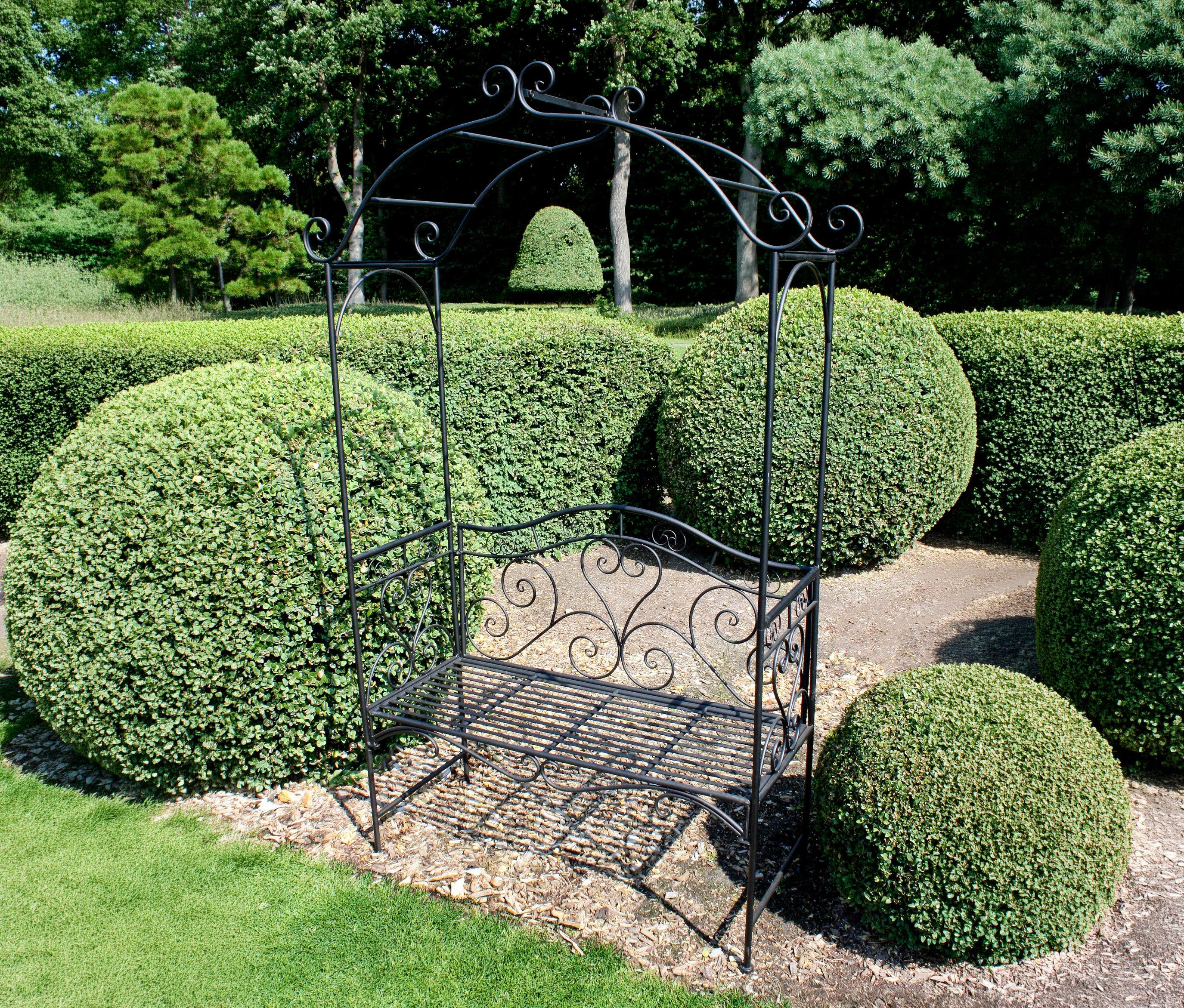 Eine schwarze Metallbank mit Rankenmuster steht unter einem Bogen in einem Garten mit geschnittenen Büschen.