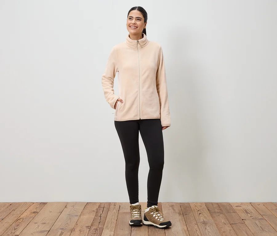Frau in beiger Fleecejacke mit Reißverschluss, schwarzen Leggings und braunen Wanderschuhen vor einer weißen Wand.