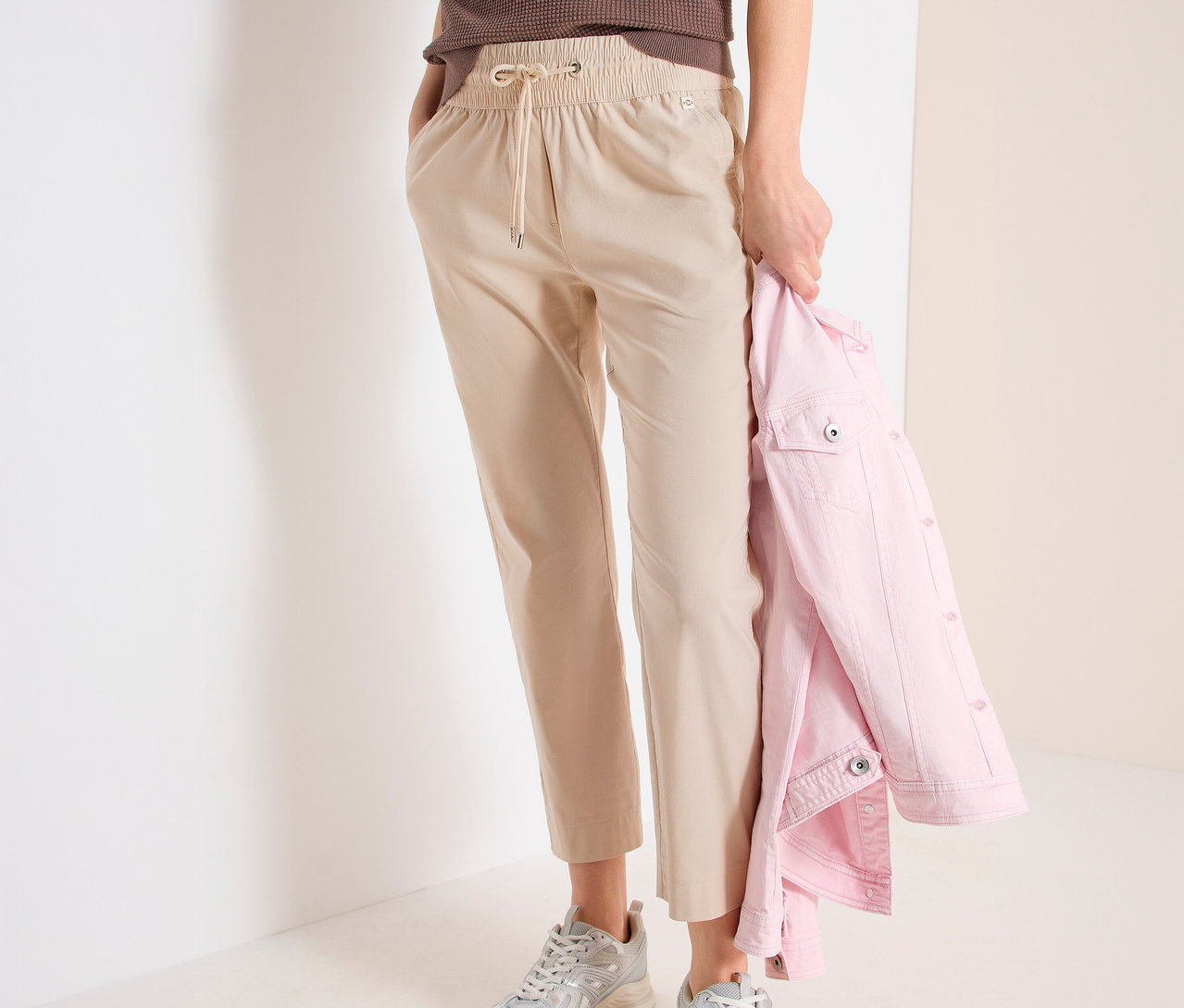 Frau mit CECIL Joggpants »Tracey«, braunem Top und silbernen Turnschuhen. Sie hält eine rosa Jacke in der Hand.