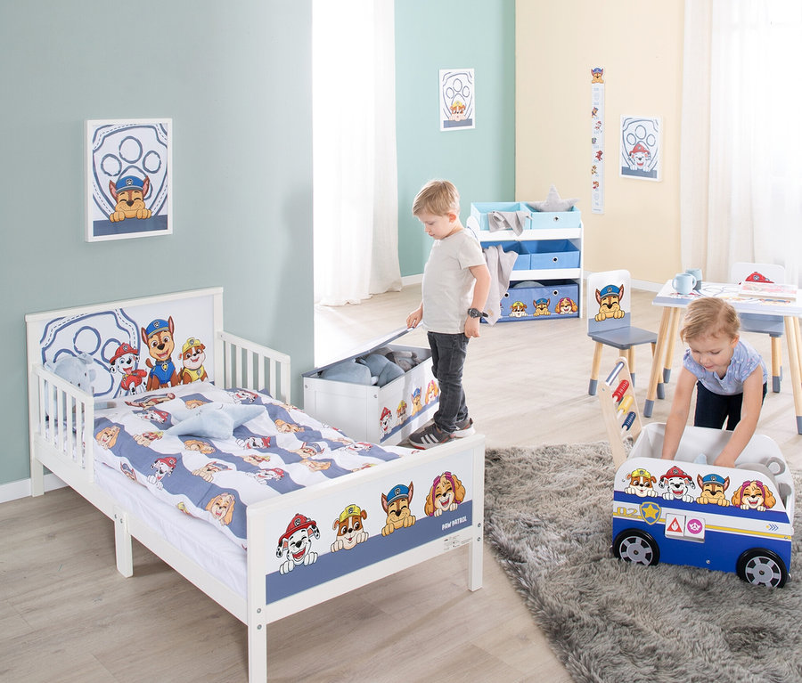 Zwei Kinder spielen in einem Zimmer mit Paw Patrol-Motiven. Ein Junge steht auf einem weißen Kinderbett mit Paw Patrol-Motiven und öffnet eine Spielzeugkiste. Ein Mädchen beugt sich über eine blaue Spielzeugkiste in Busform.