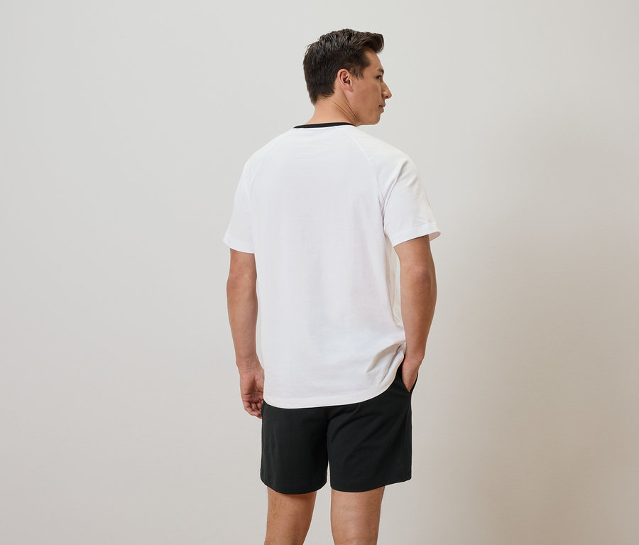 Mann von hinten in weißem T-Shirt und schwarzen Shorts, Hände in den Taschen.