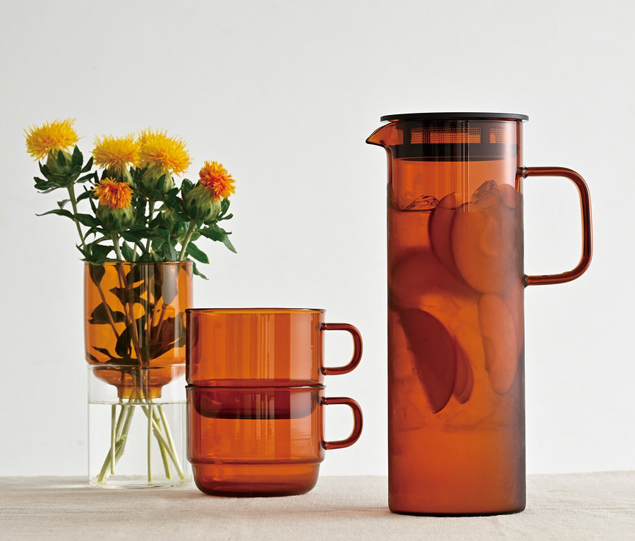 Gelbe Blumen in HARIO »Colors« Tee- und Kaffee-Set, amberfarbener Vase, zwei übereinander gestapelte Tassen und ein Krug mit Inhalt stehen auf einem Tisch.