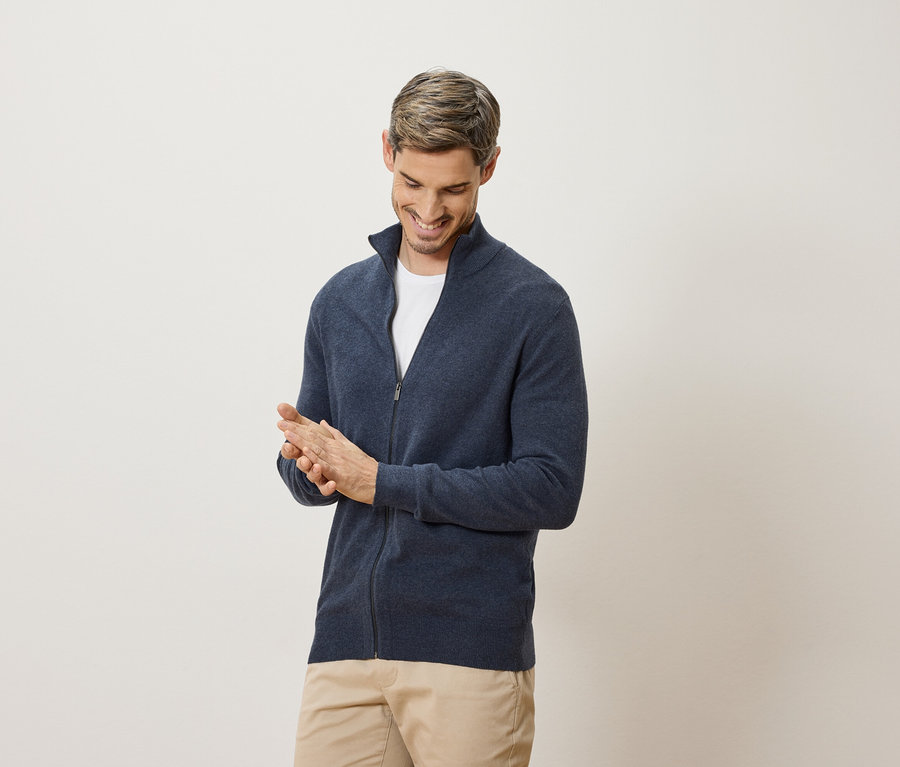 Ein Mann lächelt und reibt sich die Hände. Er trägt eine marineblaue Strickjacke und eine beige Hose.