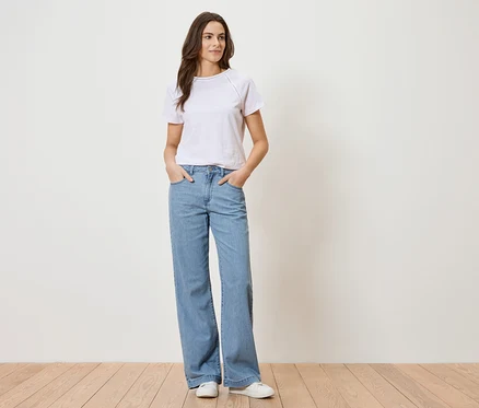 Frau mit weißem T-Shirt und blauen Jeans steht vor einer weißen Wand.