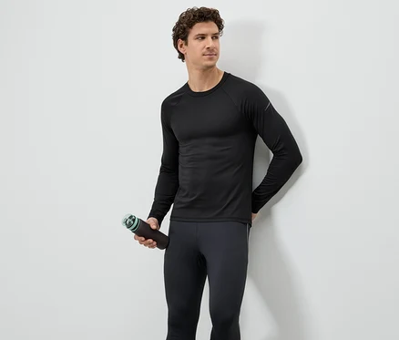 Mann in schwarzem Sportoberteil und Leggings hält eine braune Wasserflasche vor einer weißen Wand.