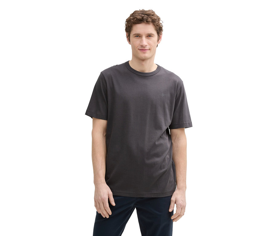 Mann mit lockigem Haar trägt ein graues T-Shirt und blaue Hosen, stehend vor einem weißen Hintergrund.