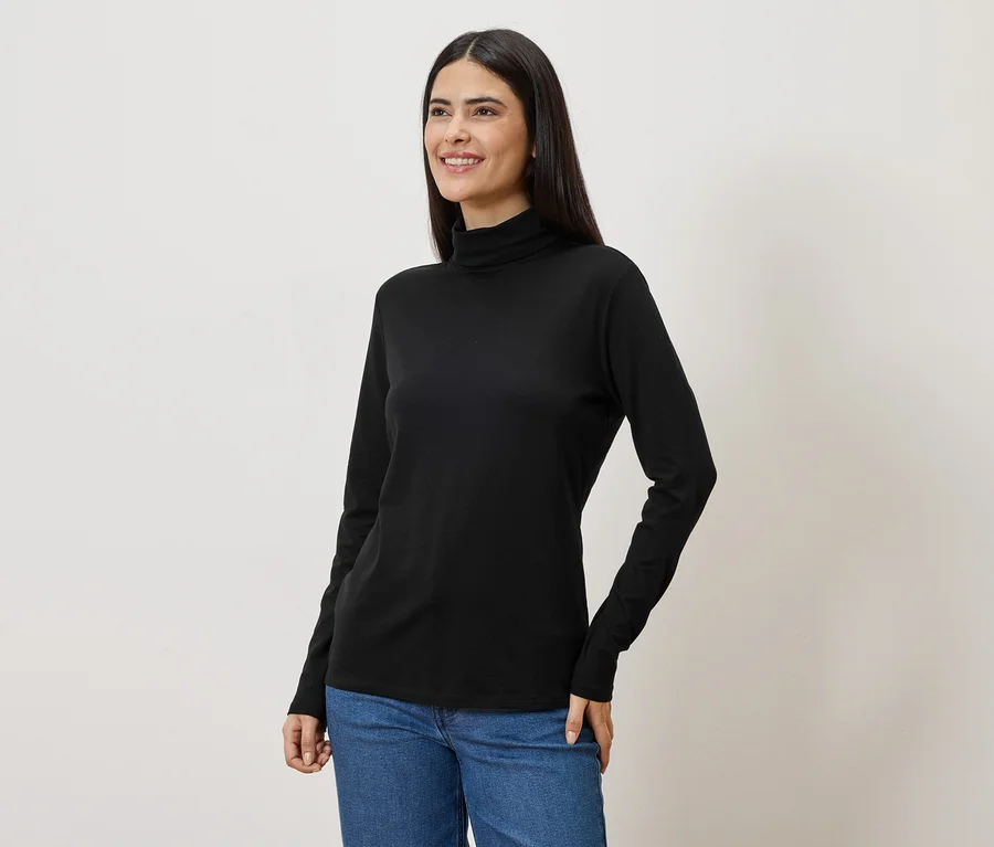 Eine Frau mit langen, dunklen Haaren trägt ein schwarzes Rollkragenshirt und Jeans.