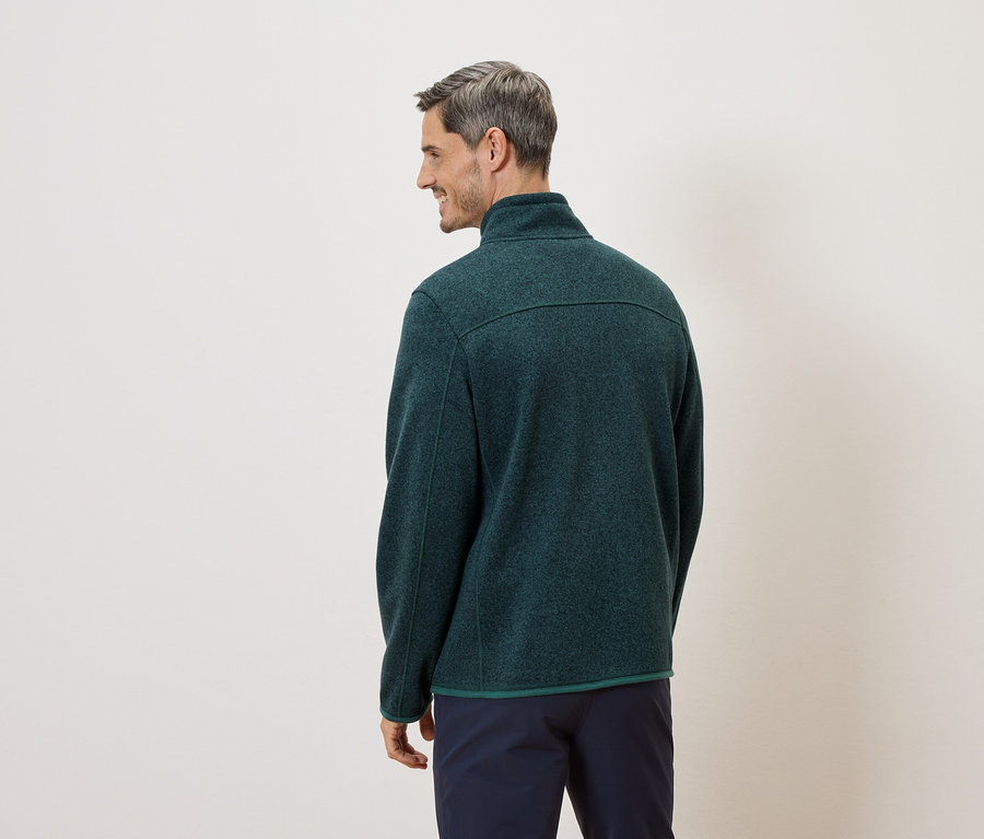 Ein Mann mit grauen Haaren, von hinten gesehen, trägt eine grüne Fleecejacke und marineblaue Hosen vor einem cremefarbenen Hintergrund.