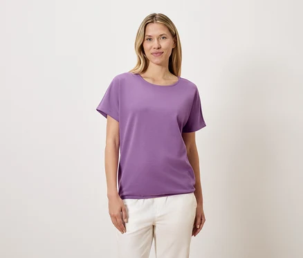 Frau mit blonden Haaren trägt ein violettes T-Shirt und weiße Hosen.